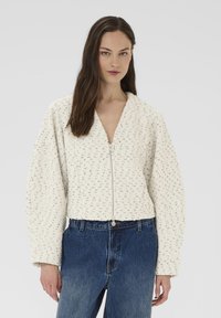 Hvid, struktureret lynlåstrøje med V-hals og vide ærmer. Kombineret med højtaljede blå denimjeans.