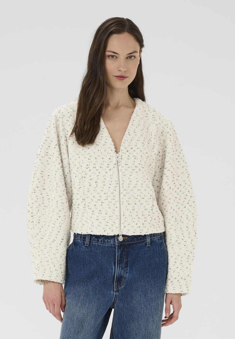 Balta, teksturēta jaka ar rāvējslēdzēju, V veida apkakli un platajiem piedurknēm. Kombinēta ar augsta vidukļa zilām džinsa biksēm.