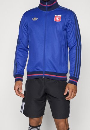 Mand iført en blå lynlåstrøje med røde og sorte striber og sorte shorts med hvide striber, stående mod en ensfarvet baggrund.