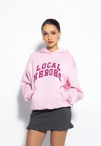 Pink hoodie featuring "LOCAL HEROES" in burgundy text, with a large front pocket, worn over a gray pinstriped skirt. Smooth texture.
