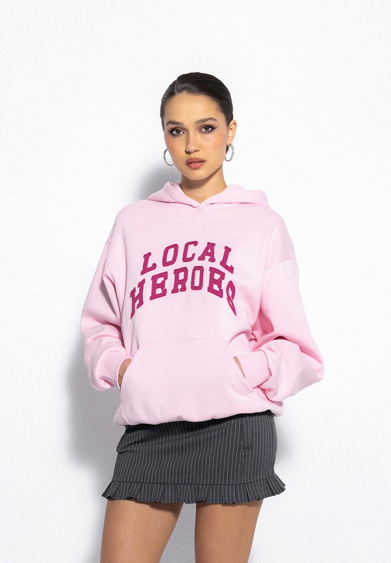 Pink hoodie featuring "LOCAL HEROES" in burgundy text, with a large front pocket, worn over a gray pinstriped skirt. Smooth texture.