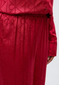 Czerwony komplet piżamowy z satynowego materiału, z detalem z guzikami i geometrycznym wzorem na całej powierzchni. Pas posiada elastyczną taśmę.