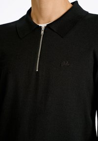Polo nero con colletto zip, realizzato in tessuto testurizzato. Presenta un logo ricamato sul lato sinistro del petto.