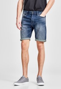 Muž v vykasaných modrých džínových šortkách, černém tričku a šedých teniskách stojící s jednou rukou v kapse na bílém pozadí.