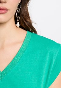 Groene V-hals top gemaakt van zachte stof met een sprankelende rand, gecombineerd met een stevige zilveren kettingoorbel met een parelaccent.