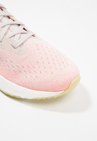 Zapatilla deportiva rosa claro con parte superior de tejido transpirable, cordones grises y una entresuela blanca. Posee textura perforada para ventilación.