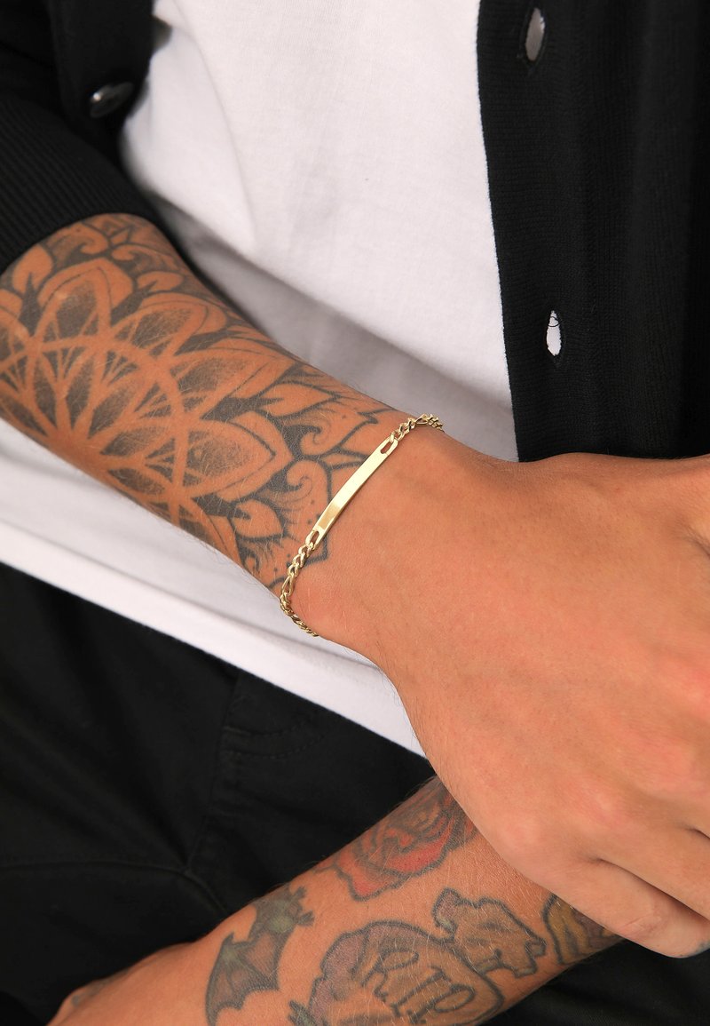 Bracciale d'oro con un design a barra piatta e catena attorcigliata, indossato su un polso adornato da tatuaggi intricati, il tutto su uno sfondo di un cardigan nero.
