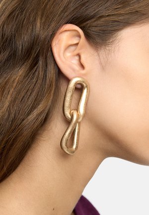 Zbliżenie ucha z błyszczącymi, złotymi, dużymi kolczykami w formie łańcucha na tle brązowych włosów i jasnej skóry.