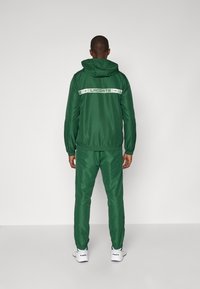 Green athletic tracksuit with a hood, featuring white "LACOSTE" branding on the back. Smooth texture, elastic cuffs, and fitted silhouette.