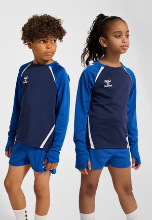 Zwei Kinder, die blaue Sportshirts und Shorts tragen, stehen vor einem einfarbigen Hintergrund und schauen in entgegengesetzte Richtungen.