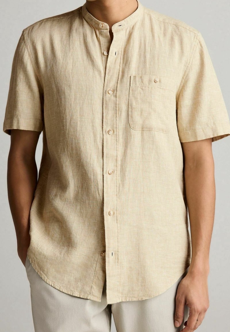 Chemise beige à manches courtes avec col, en tissu texturé, dotée d'une poche poitrine et d'une fermeture à boutons, ourlet légèrement arrondi.