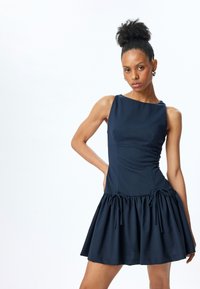 Navyblauwe jurk met een aansluitend lijfje en een uitlopende rok met ruffles en trekkoordaccenten in de taille. Gladde stoftextuur.