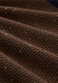 Nahaufnahme eines braunen gerippten Strickstoffs mit kleinen weißen und orangen Sprenkeln, die die Textur und sanften Falten zeigen.