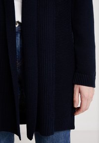 Granatowy sweter na drutach z prążkowaną fakturą, otwarty z przodu i z długimi rękawami. W dolnej części kadru widoczna ręka oraz jeansy.