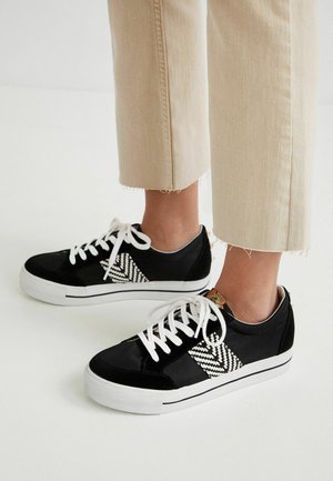 Baskets noires avec un panneau latéral texturé noir et blanc, des lacets blancs et une semelle blanche, avec un détail de design subtil au niveau du talon.
