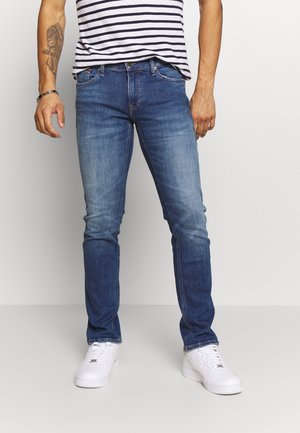 Blue denim jeans with a slim fit, featuring a faded finish, five pockets, and a zip fly. Worn with a striped shirt and white sneakers.