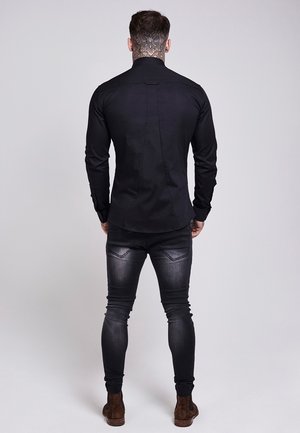 Mężczyzna z tatuażem na szyi w dopasowanej czarnej koszuli, wyblakłych czarnych obcisłych dżinsach i brązowych zamszowych butach, stojący tyłem.