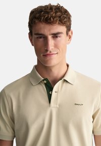 Beige polo skjorta med grön krage och två knappar, med en broderad "GANT"-logotyp på vänster sida. Slät bomullstextur.