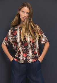 Chemise ample à motif floral beige, ornée de fleurs rouges et bleues avec des rayures verticales. Associée à un pantalon bleu foncé à coupe décontractée.