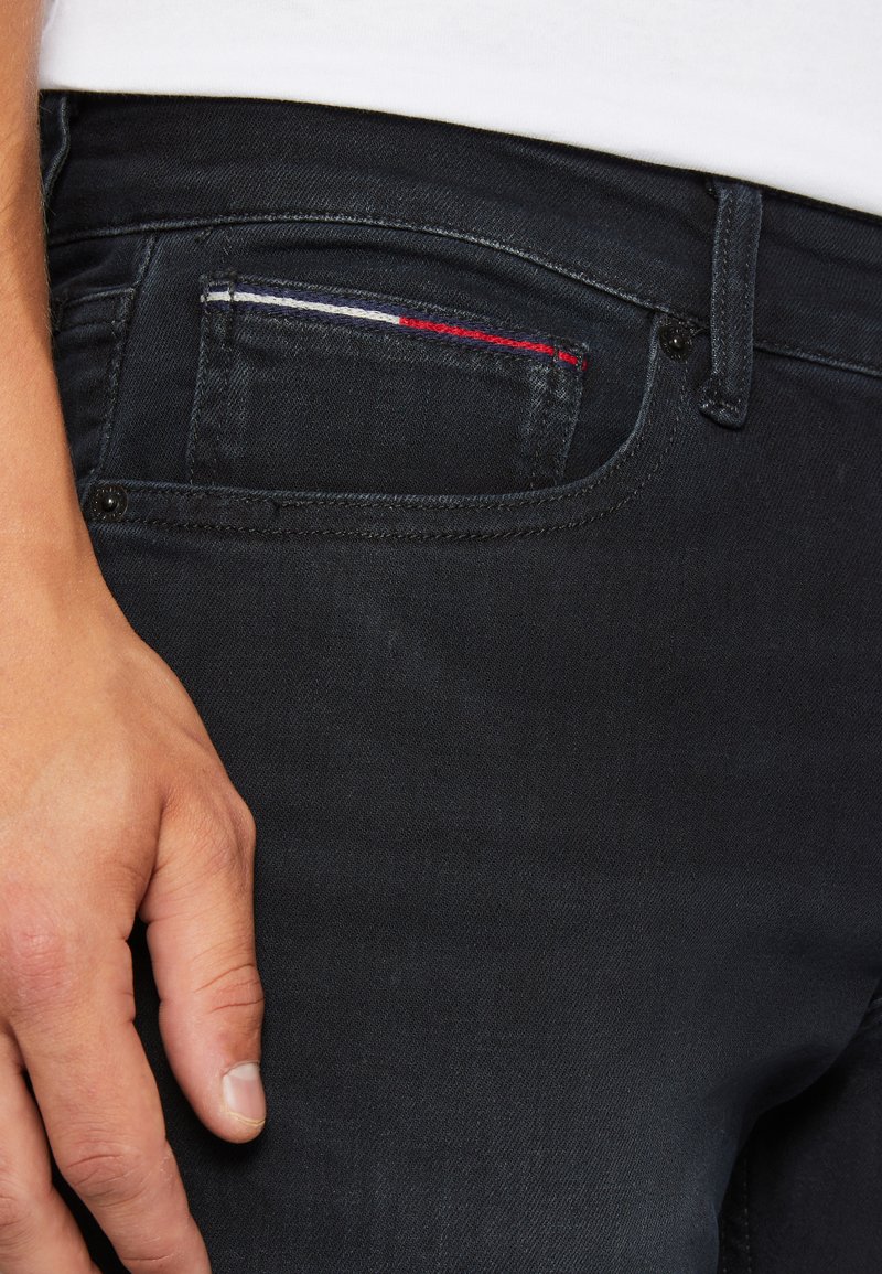 Czarne dżinsy z przodu z kieszenią, ozdobioną czerwono-biało-niebieskimi przeszyciami. Gładka tekstura i dopasowany krój. Ręka oparta na pasku.