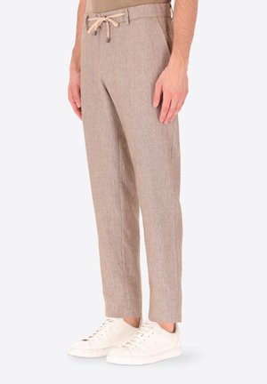 Uomo che indossa pantaloni di lino beige con coulisse e sneakers bianche, in piedi di lato su uno sfondo semplice.