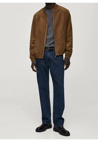 Homme portant une veste bomber en daim marron, un pull gris, un jean bleu foncé, une ceinture noire et des bottes en cuir noir, debout devant un fond uni.