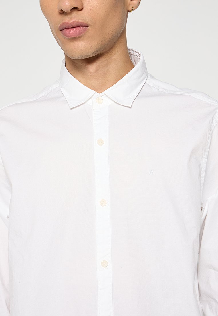 Weißes Hemd mit Knöpfen aus glattem Baumwollstoff, das einen klassischen Kragen, ein schlichtes Design und ein dezentes Logo auf der Brust aufweist.