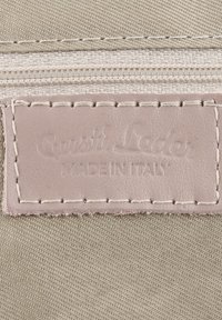 Beige kangas, jossa on teksturoidulla pinnalla vaaleanpunainen nahkainen merkki, johon on painettu "Gusti Leder" ja "MADE IN ITALY." Ompeleet näkyvät reunoilla.