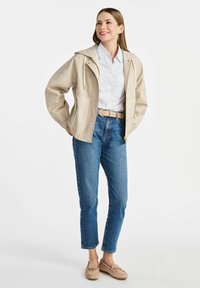 Beżowa wodoodporna kurtka z kapturem, zamkiem błyskawicznym i guzikami na zatrzask, noszona na białej i niebieskiej koszuli w paski oraz spodniach dżinsowych z wysokim stanem.