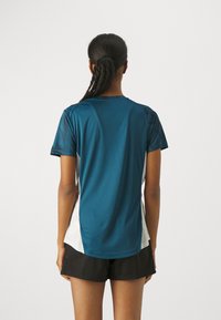 Kvinna med flätad hästsvans som bär en blå och vit t-shirt med kort ärm och svarta shorts, stående vänd bort från kameran mot en neutral bakgrund.