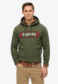 Zielona oliwkowa bluza z kapturem z grafiką z przodu, na której widnieje napis "Superdry". Posiada kieszeń kangurkę, kaptur ze sznurkiem oraz ściągacze przy rękawach.
