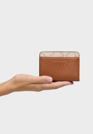 Mano sosteniendo una pequeña tarjetero de cuero marrón con el relieve "TOUS" y detalle beige con patrón en la parte superior sobre un fondo liso.