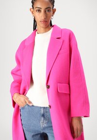 Ljusrosa ullkappa med kavajkrage, framknäppning med knappar och en enda bröstficka. Ihopparad med en krämfärgad tröja och blå jeans.