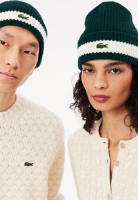 Berretti a maglia a strisce verdi e bianche abbinati a un maglione e a un cardigan di un crema texture, entrambi con un piccolo logo ricamato.