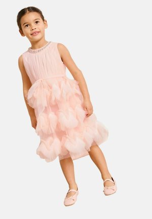 Fiatal lány ujjatlan rózsaszín ruhában, rétegzett tülllel és szív alakú díszítéssel, rózsaszín balettcipőben, egyszerű háttér előtt áll.