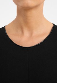 Schwarzes Kleid mit rundem Ausschnitt und feiner schwarzer Einfassung, glattem Stoff und dezentem Nahtdetail in der Mitte.