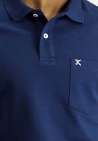 Polo shirt blu navy con colletto a coste, tre bottoni e taschino sul lato sinistro del petto con un piccolo dettaglio ricamato bianco. Tessuto morbido.