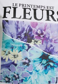 Vit tyg t-shirt med ett blommönster i nyanser av blått, lila och gult. Texten "LE PRINTEMPS EST FLEURS" står framträdande.