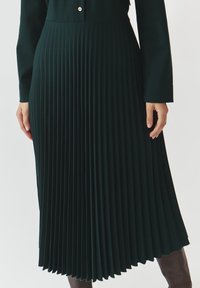 Gonna plissettata verde scuro con una finitura testurizzata. Presenta una vita aderente e un design fluido, che termina sotto il ginocchio, con dettagli a bottoni.