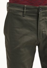 Gros plan de la taille de pantalon vert foncé avec bouton, passants de ceinture et fermeture éclair, porté avec une chemise rouge texturée.
