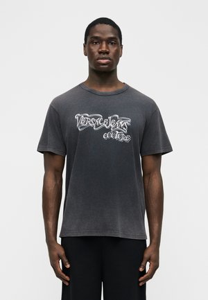Jeune homme de face portant un t-shirt noir délavé à manches courtes avec un texte graphique blanc "Versace Jeans Couture" sur le devant.