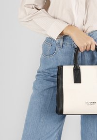 Torba płócienna z czarnymi skórzanymi akcentami, prostokątna forma i krótkie, mocne uchwyty. Na przedniej stronie znajduje się napis "CAFENOIR zrobione z miłością".