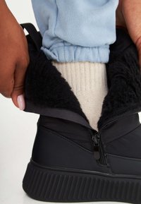 Botină neagră rezistentă la apă, cu fermoar și căptușeală din blană sintetică neagră. Talpa texturată vizibilă și pantaloni cu manșetă de culoare albastră deschisă.