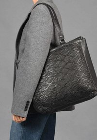 Zwarte geweven leren totebag met een glad bovenste gedeelte en een gestructureerde onderkant; heeft een stevige strap en een gestructureerde vorm.