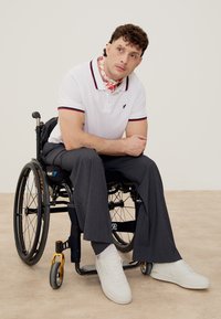 Man seated in a wheelchair wearing a white polo, gray pants, white sneakers, and a red floral neck scarf, looking thoughtfully to the side.