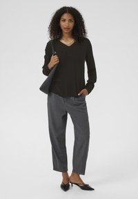 Zwarte langemouwen shirt met een v-hals en knoopdetails, gecombineerd met grijze tapered broek. Model draagt een zwarte schoudertas en heeft puntige hakken aan.