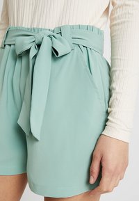 Mátově zelené kraťasy s vysokým pasem vyrobené z lehkého materiálu, s bočními kapsami a odpovídajícím páskem se mašlí.