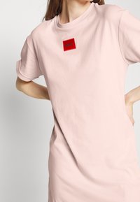 Femme portant une robe rose clair avec des manches courtes retroussées et un petit patch de logo carré rouge indiquant "HUGO" sur la poitrine.