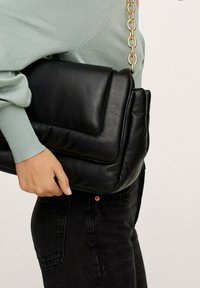 Czarna skórzana torebka o pikowanym wzorze, z złotym łańcuszkiem i wyraźnie ukształtowanym kształtem, trzymana pod jasnookiego zielonego topu z zakrzywionym dołem.