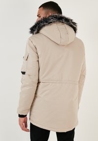 Parka bej căptușit cu glugă din blană artificială detașabilă, având un cordon la talie, buzunare laterale și detalii negre.
